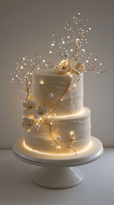 a three tiered cake with white frosting and flowers on top is lit up by fairy lights