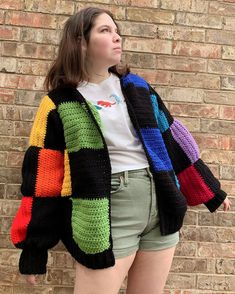 a woman standing in front of a brick wall wearing shorts and a colorful crocheted jacket