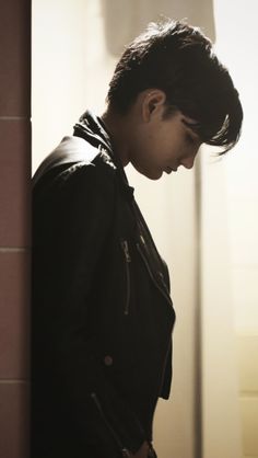 a young man standing in front of a wall looking at his cell phone while wearing a leather jacket