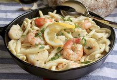 a skillet filled with pasta and shrimp on top of a striped table cloth next to bread