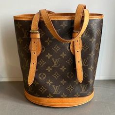 The Petit Bucket Bag From Louis Vuitton Is Made With The Brand’s Signature Brown Monogram Canvas, And Features Brass-Tone Hardware, Natural Vachetta Cowhide Leather, Two Buckled Shoulder Straps, Trim Around The Bottom Edge, And Branded Bottom Feet. The Interior Is Lined In A Grained Ivory Leather, And Features A Zippered Pocket, An Open Pocket, And A Chain To Attach Your Favourite Accessories. Condition: Very Good; Some Marking And Faint Scratching On Leather Trim, Some Wear On Base Feet, Light Wear On Hardware, Light Marking On Lining. Comes With Dustbag, Does Not Come With Pouch. Approximate Measurements: Height: 10" (25.5 Cm) Width: 8.75" (22 Cm) Depth: 6” (15 Cm) Strap Drop: 6 Brown Monogram Canvas Bucket Bag With Leather Handles, Modern Brown Monogram Canvas Bucket Bag, Brown Monogram Canvas Bucket Bag With Adjustable Strap, Elegant Brown Monogram Canvas Bucket Bag, Brown Bucket Bag With Gold-tone Hardware, Monogram Canvas, Womens Tote Bags, Leather Trims, Cowhide Leather