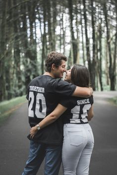 a man and woman standing next to each other in front of some trees with their arms around each other