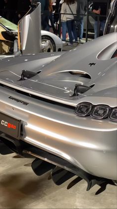 the rear end of a silver sports car on display at an auto show with people standing around