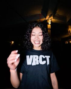 a woman with curly hair is smiling and holding something in her hand while wearing a vcct t - shirt