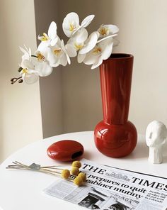 a red vase with white flowers in it sitting on a table next to a newspaper