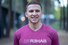 a man in a purple shirt smiles at the camera with trees in the back ground