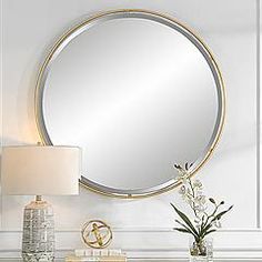 a round mirror on the wall above a table with flowers and a lamp next to it