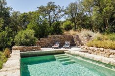 an outdoor swimming pool surrounded by stone walls and greenery, with two lawn chairs on the side