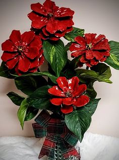 red flowers are in a glass vase on a white surface with green leaves and plaid ribbon