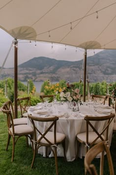 an outdoor table set up with white linens and wooden chairs, under a large umbrella