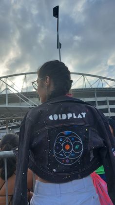 a woman wearing a jacket with the word godplay painted on it, standing in front of a stadium