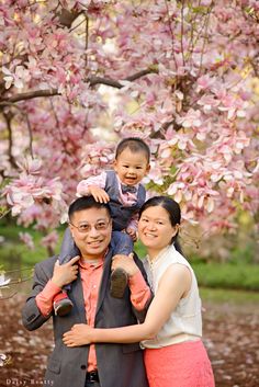 a man and woman holding a baby in front of a tree with pink flowers on it