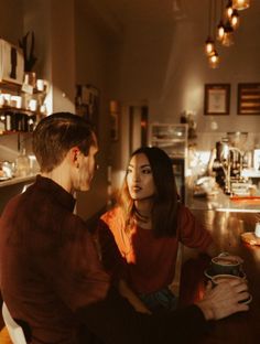 a man and woman sitting at a bar talking to each other with coffee cups in front of them