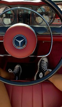 the interior of an old car with red leather seats and steering wheel, while someone's feet are on the floor