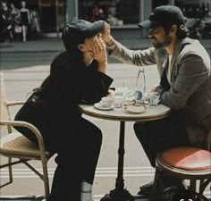 two people sitting at a table with cups and saucers in their hands, one is touching the other's face