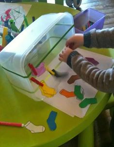 a child is playing with construction paper and scissors on a table in front of a plastic container