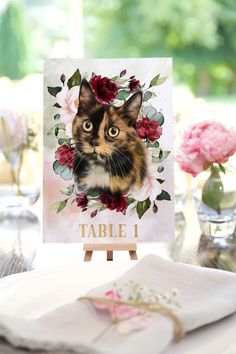 a card with a cat's face on it and flowers in the background, sitting on a table