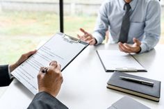 two men sitting at a table with papers in front of them and one man holding a clipboard