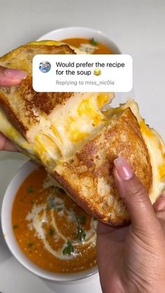 someone is holding up a grilled cheese sandwich with soup in the bowl behind it