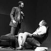 two men standing on stage with one sitting in a chair