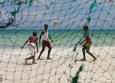 three men playing soccer on the beach with net in foreground and ocean in background