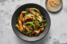 a black bowl filled with stir fry vegetables and sesame seeds next to a spoon on a gray surface