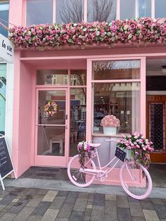 a pink bicycle parked in front of a storefront with flowers on the window and outside