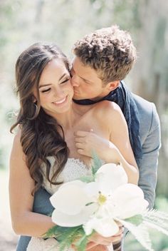 a bride and groom embracing each other in front of some trees with white flowers on their bouquet