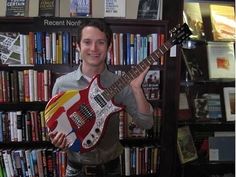 a man holding a guitar in front of a bookshelf