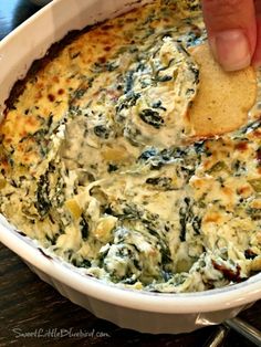 a hand dipping a tortilla chip into a bowl of spinach and artichoke dip