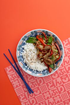 a blue and white bowl filled with rice and vegetables next to chopsticks on an orange background