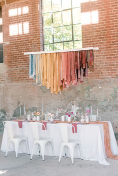 a long table is set up with white chairs and colorful scarves hanging from the ceiling