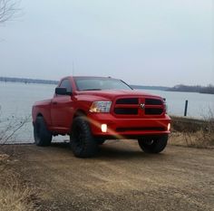 a red truck parked on top of a dirt road next to a body of water