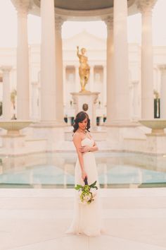 a woman standing in front of a fountain wearing a white dress and holding a bouquet