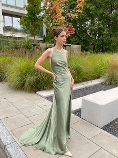 a woman in a long green dress standing on the sidewalk near some bushes and trees