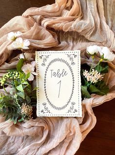 a table number sign with flowers and greenery on top of a piece of cloth