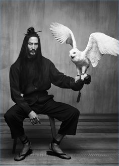 a man with long hair sitting on a chair holding an owl in front of his face