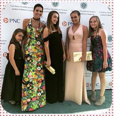 four women standing next to each other in front of a white wall and green carpet