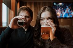 a man and woman sitting next to each other drinking from beer mugs in front of a tv