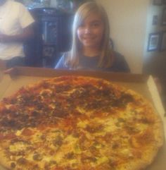 a woman sitting in front of a large pizza on top of a cardboard box with toppings