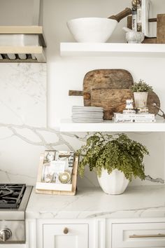 a white kitchen with marble counter tops and shelves filled with books, plants and cookbooks