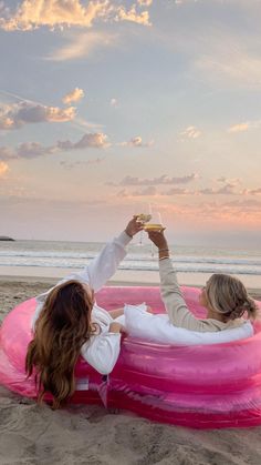 two women are sitting on an inflatable pool at the beach and drinking wine