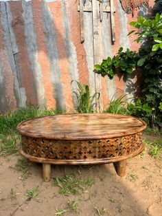 a round wooden table sitting on top of a dirt ground next to a green bush