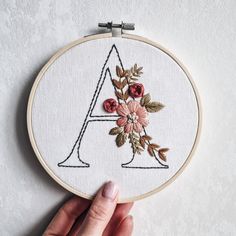 a hand is holding up a embroidery hoop with flowers on it and the letter a