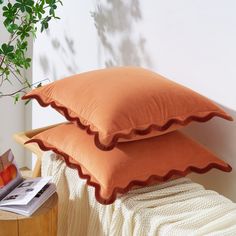 two orange pillows sitting on top of a bed next to a wooden table with a plant