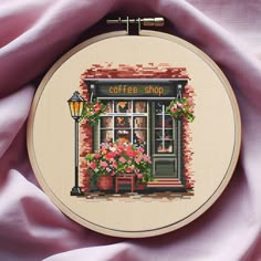 a cross stitch pattern with a coffee shop window and flowers on the outside, in front of a pink fabric