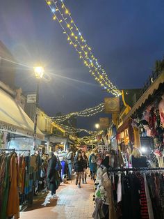 people are walking through an outdoor market with lights strung over the buildings and shops on both sides