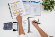 a person is writing on a notebook with a cell phone and plant in the background