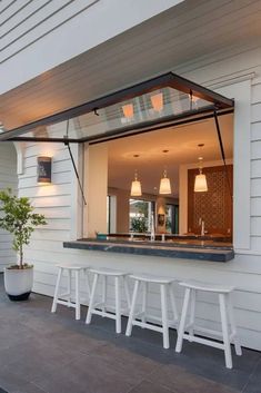 an open kitchen window on the side of a white house with stools and a potted plant