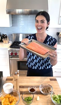 a woman holding up a tray of salmon on top of a kitchen counter next to bowls of vegetables and seasonings